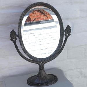 May include: Oval, antique-style vanity mirror with a dark, metallic finish. The mirror has a decorative frame and is supported by a pedestal base. The mirror's design includes floral accents and curved support arms.
