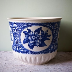 Op de afbeelding: Een grote, witte keramische bloempot met een blauw bloemmotief. De bloempot heeft een prominente blauwe bloem in een ovale lijst, omringd door ingewikkelde blauwe patronen. De rand en de basis zijn wit.