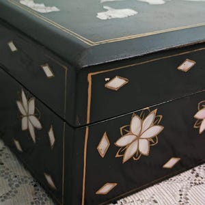 May include: A rectangular black box featuring inlaid mother-of-pearl floral motifs and diamond-shaped accents. The lid and sides are decorated with thin gold-colored lines that frame the edges, highlighting the dark finish and iridescent shell inlays.