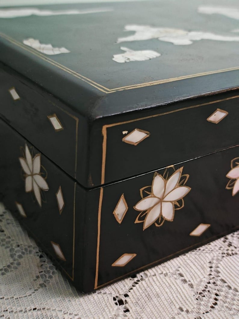 May include: A rectangular black box featuring inlaid mother-of-pearl floral motifs and diamond-shaped accents. The lid and sides are decorated with thin gold-colored lines that frame the edges, highlighting the dark finish and iridescent shell inlays.