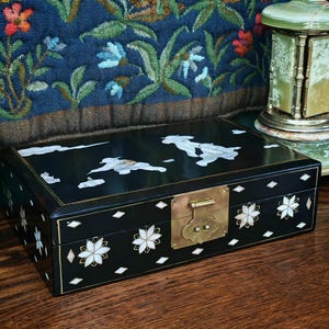 May include: A rectangular black lacquered box featuring intricate mother-of-pearl inlay designs of figures and clouds on the lid, with floral patterns and diamond shapes on the sides, secured by a central brass latch and hinge mechanism.