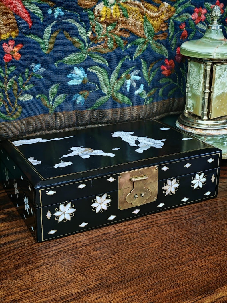 May include: A rectangular black lacquered box featuring intricate mother-of-pearl inlay designs of figures and clouds on the lid, with floral patterns and diamond shapes on the sides, secured by a central brass latch and hinge mechanism.