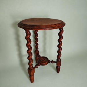 Op de afbeelding: Een ronde houten bijzettafel met een donkerbruine afwerking. De tafel heeft drie poten met een opvallend spiraalvormig ontwerp, verbonden door een centraal cirkelvormig steunstuk nabij de basis. Het tafelblad is glad met een licht opstaande rand.