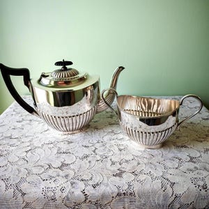 Puede incluir: Un juego de té plateado con tetera y crema. La tetera tiene un asa negra y una tapa decorativa. La crema tiene dos asas. Ambas piezas tienen un diseño acanalado y están colocadas sobre un mantel de encaje blanco.