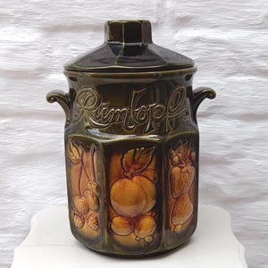 May include: A vintage ceramic jar with a dark green glaze and a matching lid. The jar features embossed fruit designs and the word "Rimtopf" in raised lettering. It has two small handles and is likely used for storage.
