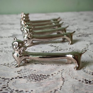 May include: A set of four silver-toned metal cat-shaped chopstick rests. Each piece features a feline head with a small bow tie and an elongated, slender body that extends to a pointed tail, designed to hold utensils in a horizontal position.
