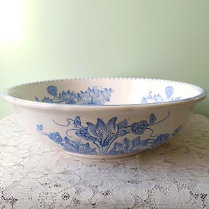 May include: A white ceramic bowl with a blue floral pattern. The bowl has a wide, shallow shape with a scalloped rim. The blue floral pattern includes stylized flowers and leaves, creating a vintage aesthetic.