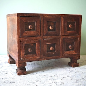 May include: A small, dark brown wooden chest of drawers with six square drawers. Each drawer has a raised square panel and a round knob. The chest sits on four short, carved legs. The wood has a warm, aged appearance.