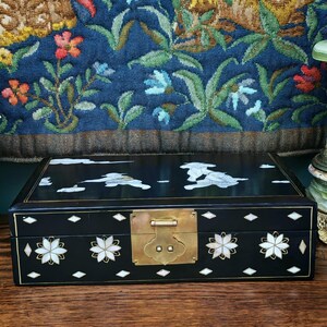 May include: A rectangular black lacquered box featuring intricate mother-of-pearl inlay designs of clouds and figures on the lid. The front panel displays floral motifs and diamond shapes with a central brass latch mechanism for closure.