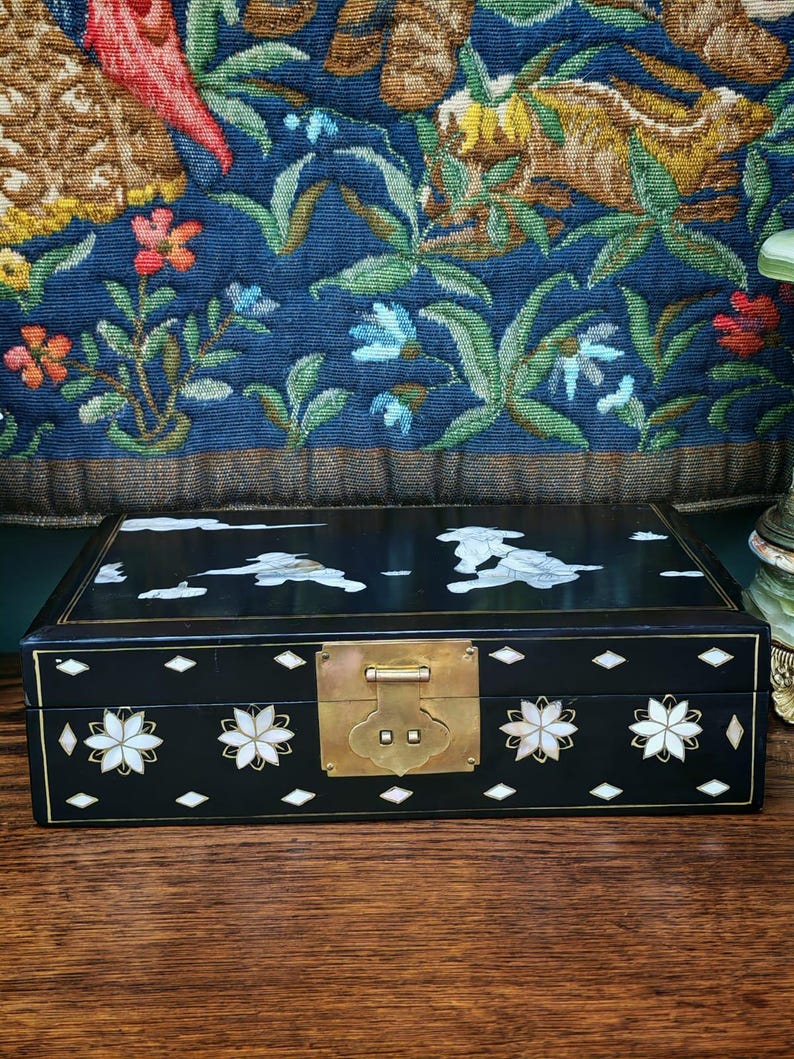 May include: A rectangular black lacquered box featuring intricate mother-of-pearl inlay designs of clouds and figures on the lid. The front panel displays floral motifs and diamond shapes with a central brass latch mechanism for closure.