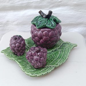 Op de afbeelding: Een keramische druiventhema set met een pot met deksel, twee zoutvaatjes en een blad-vormige schaal. De pot en zoutvaatjes zijn paars, met groene bladdetails. De schaal is groen. De set is ontworpen om op een druiventros te lijken.