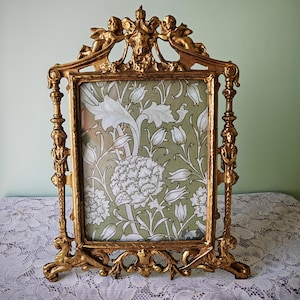 May include: Ornate gold-colored picture frame with cherub and floral details. The frame holds a green and white floral patterned print. The frame has a decorative base and is standing on a white lace tablecloth.
