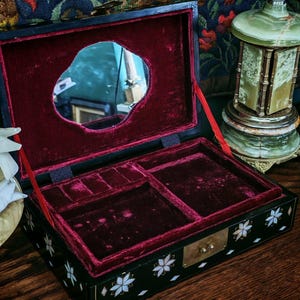 May include: A black rectangular jewelry box with a hinged lid featuring an interior mirror and deep red velvet lining. The exterior is decorated with white floral mother-of-pearl inlays and small diamond shapes, finished with a brass latch closure.