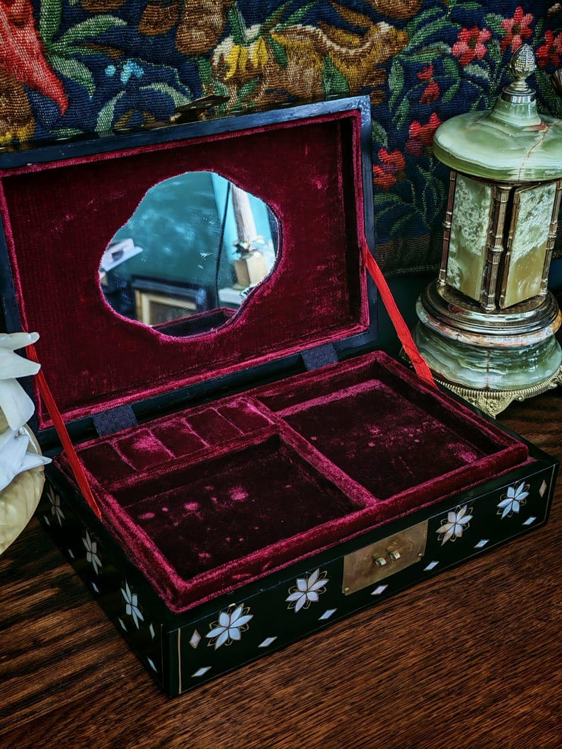 May include: A black rectangular jewelry box with a hinged lid featuring an interior mirror and deep red velvet lining. The exterior is decorated with white floral mother-of-pearl inlays and small diamond shapes, finished with a brass latch closure.