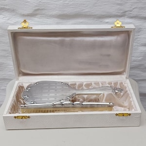 May include: A vintage vanity set in a white box with gold-colored clasps. The set includes a silver-toned hand mirror and comb. The mirror has a decorative handle and a patterned back. The comb has a light-colored handle and fine teeth. The box is lined with a satin fabric.