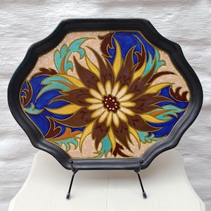 May include: A decorative black tray with a floral design. The tray features a central flower with brown, yellow, and white petals, surrounded by blue and teal accents. The tray is supported by a black metal stand.