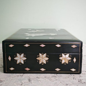 May include: A rectangular black lacquered box featuring intricate mother-of-pearl inlay. The front panel displays three floral motifs surrounded by small diamond-shaped accents. The lid shows additional inlaid figures and cloud-like shapes.