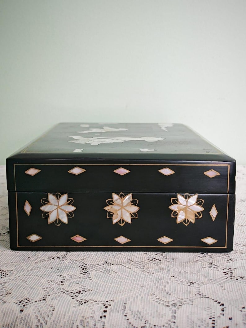 May include: A rectangular black lacquered box featuring intricate mother-of-pearl inlay. The front panel displays three floral motifs surrounded by small diamond-shaped accents. The lid shows additional inlaid figures and cloud-like shapes.