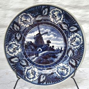 May include: A blue and white decorative plate with a windmill scene. The plate has a floral border with white and blue flowers. The plate is displayed on a black metal stand. The plate's diameter is approximately 25 cm.