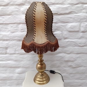May include: Vintage table lamp with a brass base and a unique lampshade. The lampshade features a woven brown pattern, tan center, and brown fringe. The lamp is on a white surface against a white brick wall.