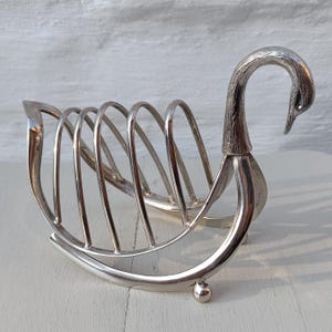 May include: A silver-colored swan-shaped toast rack. The rack features a curved base with a small ball foot on each end. The swan's neck forms a handle, and the body has vertical slots to hold toast slices. A decorative kitchen item.