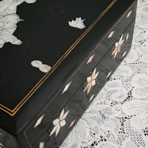 May include: A rectangular black lacquered box featuring intricate mother-of-pearl inlay. The lid displays figures and a leaf motif, while the sides are decorated with floral patterns and diamond-shaped accents, all framed by thin gold-colored lines.