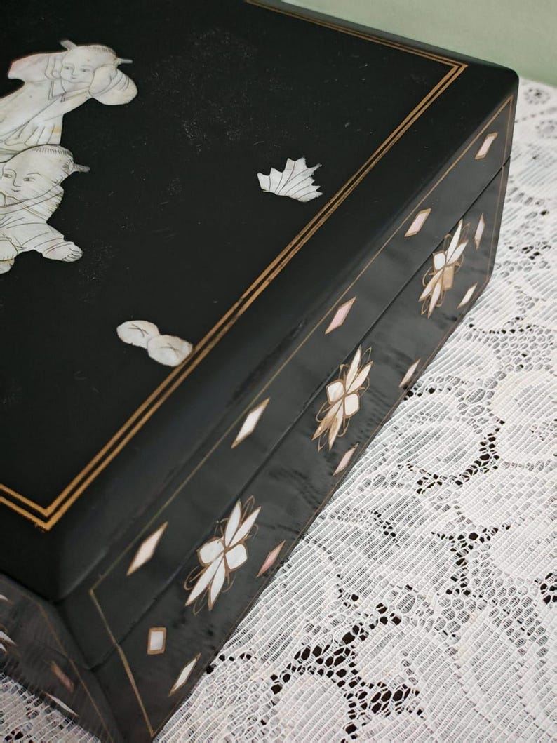 May include: A rectangular black lacquered box featuring intricate mother-of-pearl inlay. The lid displays figures and a leaf motif, while the sides are decorated with floral patterns and diamond-shaped accents, all framed by thin gold-colored lines.