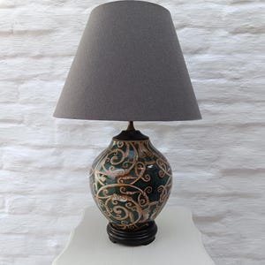 Könnte beinhalten: Eine Tischlampe mit einem konischen grauen Stoffschirm und einem abgerundeten Keramikfuß. Der Fuß zeigt einen dunkelgrünen Hintergrund mit komplexen, geschwungenen hellbraunen und cremefarbenen Rankenmustern auf einem dunkelbraunen runden Sockel.