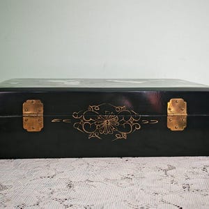 May include: A rectangular black lacquered box featuring a smooth finish, two brass-colored metal hinges, and a delicate gold-colored floral line drawing centered on the front panel between the hinges, showcasing a polished and dark exterior.