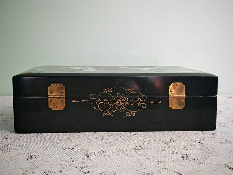 May include: A rectangular black lacquered box featuring a smooth finish, two brass-colored metal hinges, and a delicate gold-colored floral line drawing centered on the front panel between the hinges, showcasing a polished and dark exterior.