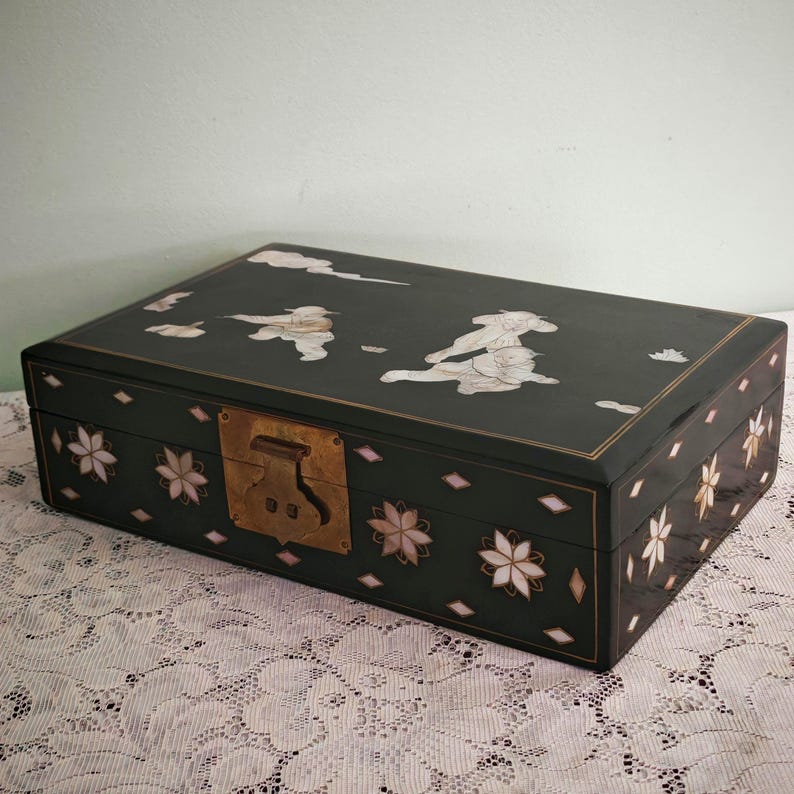 May include: A rectangular black lacquered box featuring mother-of-pearl inlay depicting figures in motion on the lid. The sides are decorated with floral motifs and diamond shapes, and the front includes a central brass latch mechanism.