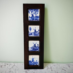 Può includere: Una placca da parete verticale in legno marrone scuro con quattro piastrelle in ceramica quadrate disposte in colonna. Ogni piastrella bianca presenta un'illustrazione a inchiostro blu di un paesaggio con mulino a vento, alberi e acqua.