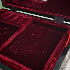 May include: A black jewelry box with a hinged lid featuring a mirror on the interior. The interior is lined with deep red velvet, including a large open compartment and a smaller section with ring slots. The exterior has decorative floral inlays.