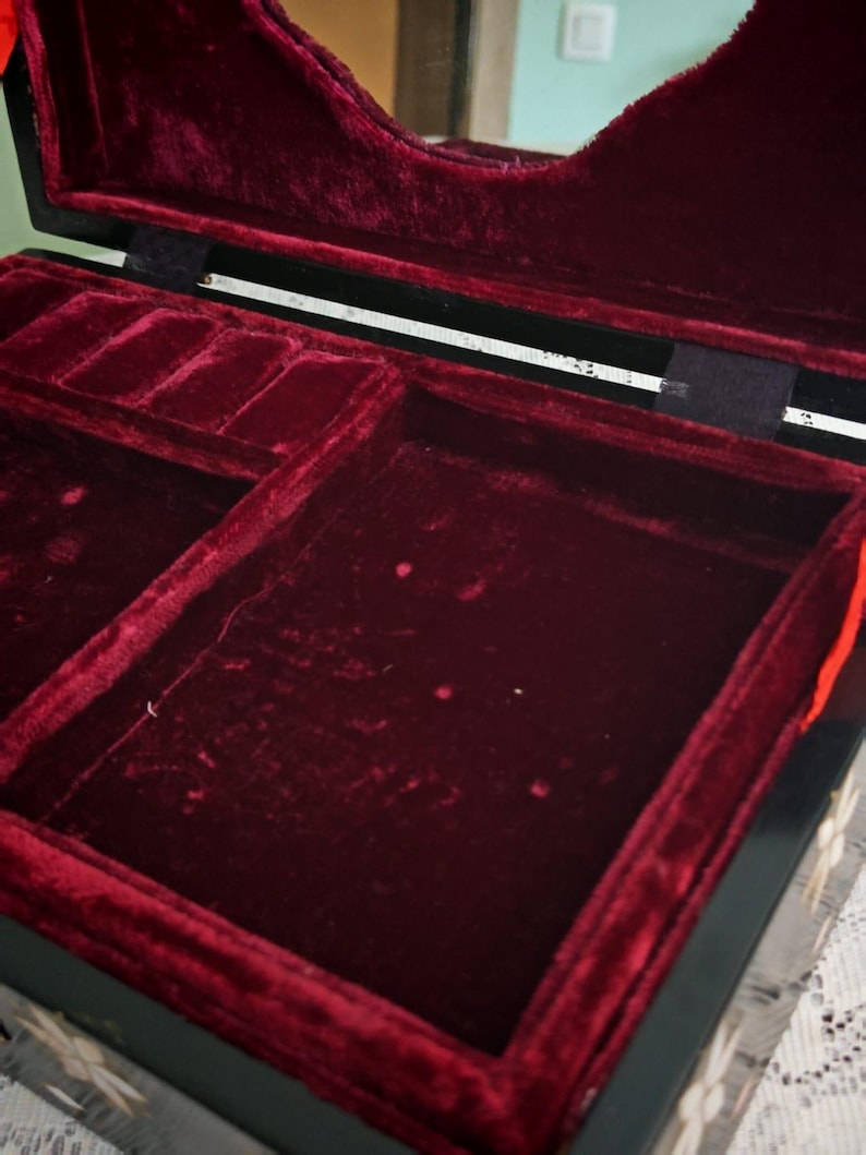 May include: A black jewelry box with a hinged lid featuring a mirror on the interior. The interior is lined with deep red velvet, including a large open compartment and a smaller section with ring slots. The exterior has decorative floral inlays.