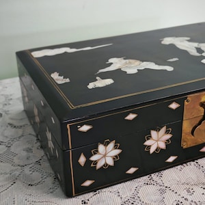 May include: A rectangular black lacquered box featuring inlaid mother-of-pearl floral motifs and cloud patterns on the lid. The front panel displays flower designs and small diamond shapes, accented by a brass latch mechanism on the right side.