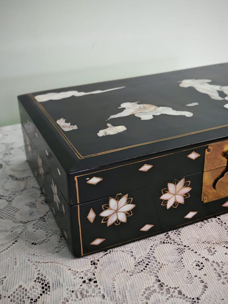 May include: A rectangular black lacquered box featuring inlaid mother-of-pearl floral motifs and cloud patterns on the lid. The front panel displays flower designs and small diamond shapes, accented by a brass latch mechanism on the right side.