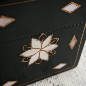 May include: A black rectangular box featuring a central white floral inlay made of shell segments, accented with gold-colored outlines. The surface is decorated with small, white diamond-shaped shell inlays bordered by gold lines.
