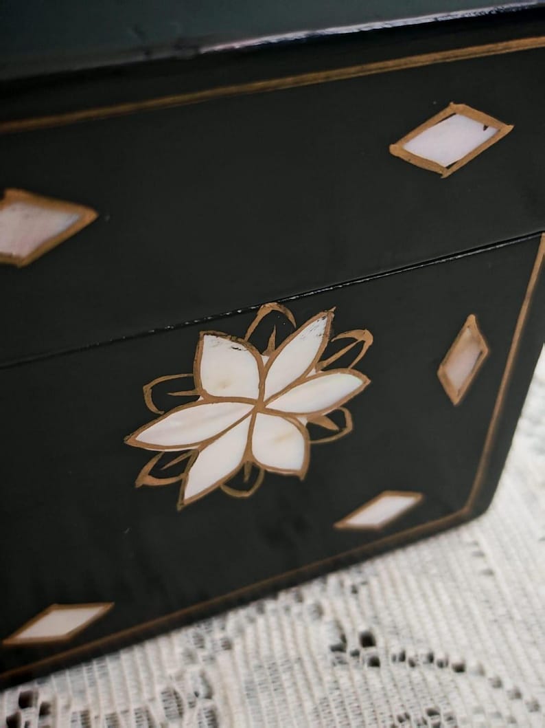 May include: A black rectangular box featuring a central white floral inlay made of shell segments, accented with gold-colored outlines. The surface is decorated with small, white diamond-shaped shell inlays bordered by gold lines.
