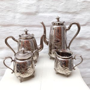 Op de afbeelding: Een zilveren theeservies met sierlijke bloemen- en bladgravures. De set bevat een theepot, een melkkan, een suikerpot en een kleinere theepot. Elk stuk heeft een deksel en staat op kleine voetjes.