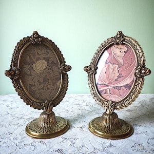 May include: Two oval-shaped brass picture frames with ornate, embossed metal borders and pedestal bases. The left frame contains a dark brown floral print, while the right frame features a pink and brown leaf pattern under clear glass.