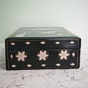 May include: A rectangular black box featuring white mother-of-pearl inlay designs. The front panel displays three floral motifs surrounded by small diamond-shaped accents. The lid also features white inlay patterns, and thin gold lines outline the edges.