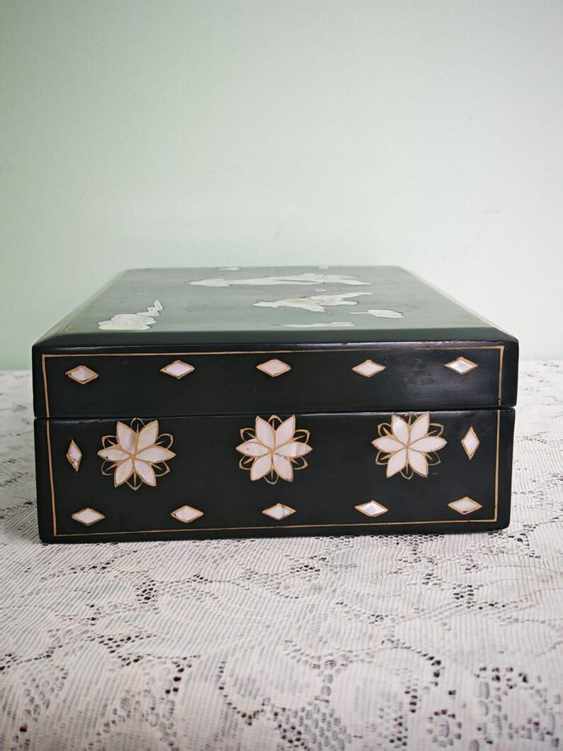 May include: A rectangular black box featuring white mother-of-pearl inlay designs. The front panel displays three floral motifs surrounded by small diamond-shaped accents. The lid also features white inlay patterns, and thin gold lines outline the edges.