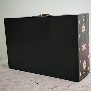 May include: A rectangular black box with a smooth finish and a metallic latch on the top edge. The side panel features a decorative pattern of white and gold floral motifs interspersed with small diamond shapes, creating a distinct visual contrast.