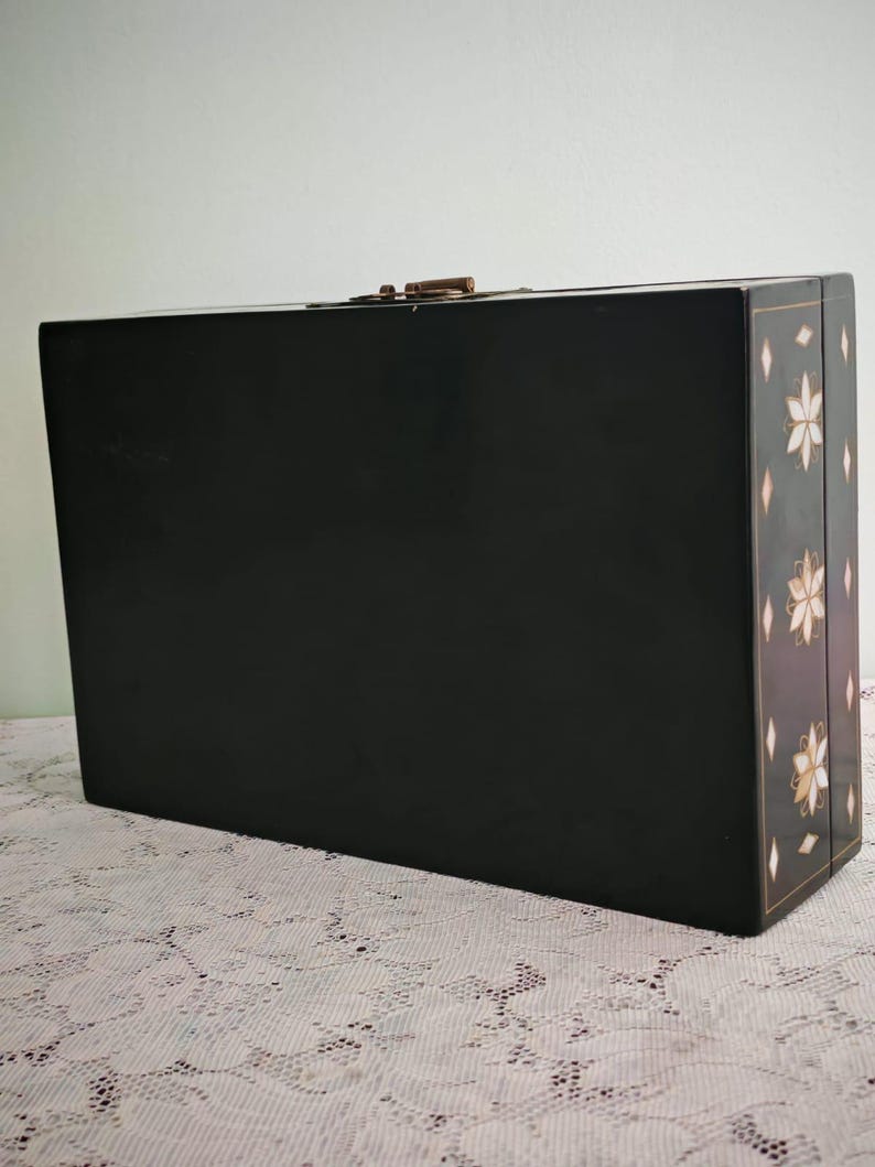 May include: A rectangular black box with a smooth finish and a metallic latch on the top edge. The side panel features a decorative pattern of white and gold floral motifs interspersed with small diamond shapes, creating a distinct visual contrast.