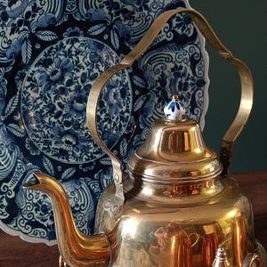 Puede incluir: Una tetera de latón pulido con un pico curvo y un asa, junto a un plato decorativo azul y blanco. La tetera tiene un pequeño pomo decorativo en la tapa. Un recipiente de cobre está en primer plano.