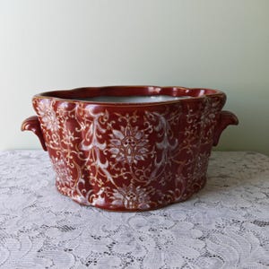 Pode incluir: Uma taça de cerâmica vermelho-escura com borda recortada e duas alças laterais. O exterior apresenta um padrão floral branco repetitivo com contornos dourados. O interior da taça é branco e a forma geral é oval e larga.