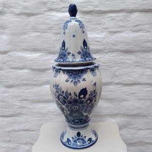 May include: A white and blue ceramic vase with a lid. The vase features a floral design in shades of blue. The vase is approximately 30 cm tall.