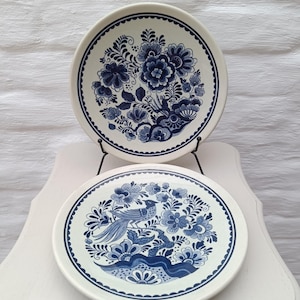 Puede incluir: Dos platos de cerámica blancos con diseños florales y de pájaros azules. Los platos tienen un borde festoneado y un borde azul. Uno presenta un patrón floral, y el otro tiene un diseño de pájaro y flores.