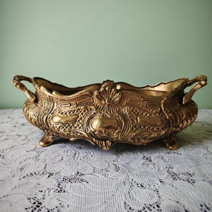 May include: An elongated, oval-shaped brass bowl featuring ornate, raised relief detailing, a scalloped rim, and two side handles. The vessel is supported by four small, decorative feet and displays a polished, metallic gold-toned finish.