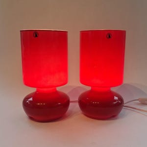 Op de afbeelding: Een paar felrode glazen tafellampen met cilindrische kappen en afgeronde voetstukken. Elke lamp heeft een kleine ronde sticker bij de bovenrand. De lampen geven een helder rood licht en zijn voorzien van witte elektriciteitssnoeren.